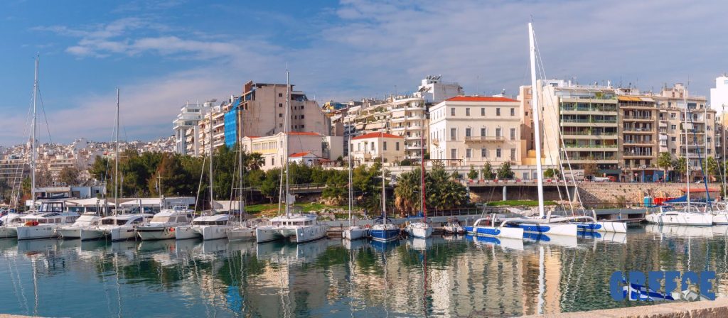 A view of a marina filled with sailboats and catamarans docked in the calm water, with a coastal city skyline of buildings and palm trees in the background under a blue sky with clouds. A logo for "GREECE" is in the bottom right corner.