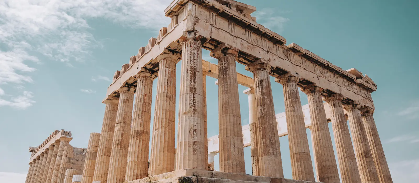 Low-angle photograph of the ancient Greek Parthenon temple on the Acropolis in Athens, showing its weathered marble columns and entablature against a light blue sky with some clouds.