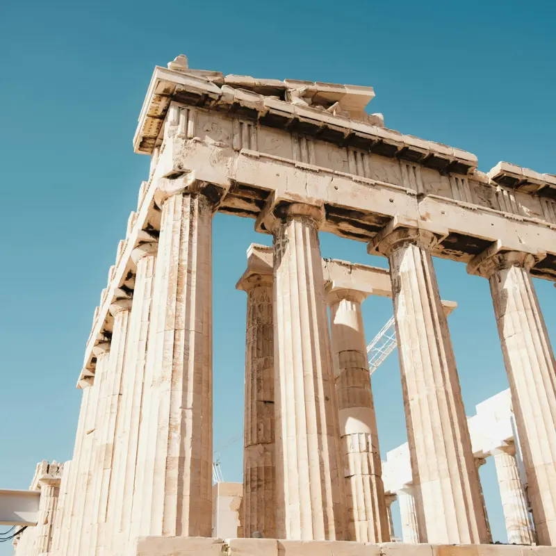 A low-angle view of the ancient marble columns and architectural ruins of the Parthenon temple under a clear blue sky in Athens, Greece.